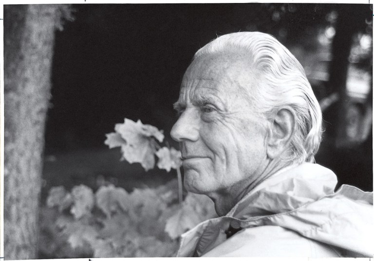 A black-and-white profile portrait shows an elderly man with slicked-back white hair looking toward the left side of the frame. Bright daylight highlights his face and light-colored jacket collar, contrasting against a dark, shaded background featuring a tree trunk and out-of-focus foliage.