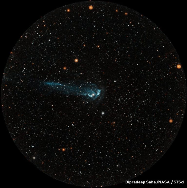 A circular image shows a bright teal, comet-like streak streaming horizontally across a pitch-black field filled with countless tiny stars. The teal streak features a brighter, compact head near the center of the circle, trailing off into a wispy blue-green tail toward the left. The surrounding dark background is dotted with mostly white and warm orange pinpricks of light, creating high contrast with the central glowing shape.