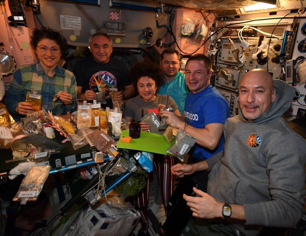 An image of Expedition 61 crew members gathered around a table with plastic food packets. The packets contain their thanksgiving meal.