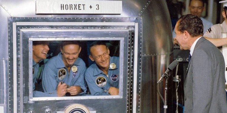 A color photograph shows a man in a grey suit standing on the right, speaking into a microphone facing a large, shiny metallic container labeled "HORNET + 3." Inside the container, three smiling men in blue jumpsuits look out through a large rectangular glass window adorned with a circular presidential seal. The indoor scene is brightly lit, showcasing the gleam of the silver metal structure against a dark blue floor.