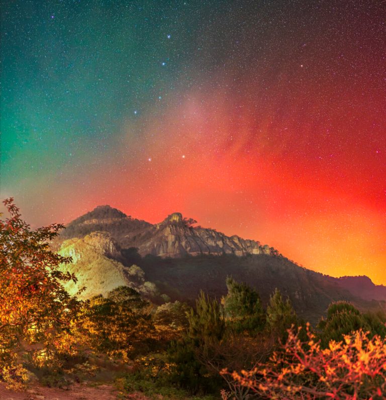 A wide night landscape view shows rocky, tree-lined mountains stretched across the center with foliage illuminated by warm yellow-orange light in the foreground. Above the dark ridge, the night sky transitions from teal green on the upper left to a vivid, glowing crimson and orange hue filling the right side, all filled with a dense field of tiny stars. A vertical line of brighter white stars stands out clearly against the colorful sky backdrop.