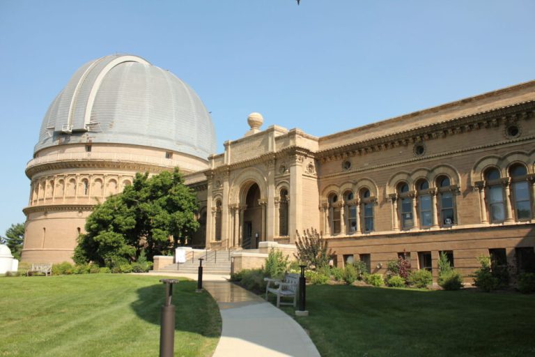 A large, tan stone building with intricate ornate detailing spans the frame under a clear, pale blue sky. On the left stands a prominent circular structure topped with a massive silver-grey dome, connected to an elongated wing featuring arched windows and a central entrance steps leading up from a paved walkway. Well-manicured green lawns, bushes, and a white bench sit in the foreground under bright, direct sunlight.