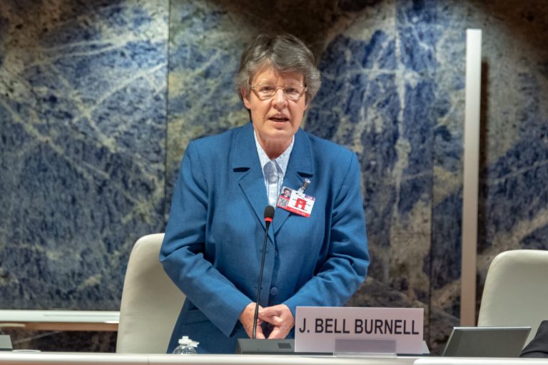 A color photograph shows a woman with short grey hair and glasses speaking from behind a long white desk, positioned in the center of the frame. She wears a blue blazer over a light-colored collared shirt, with an identification badge pinned to her lapel, and stands before a small desk microphone and a nameplate reading "J. BELL BURNELL." The setting is brightly lit, featured against a background panel with a textured, mottled blue and grey stone pattern.