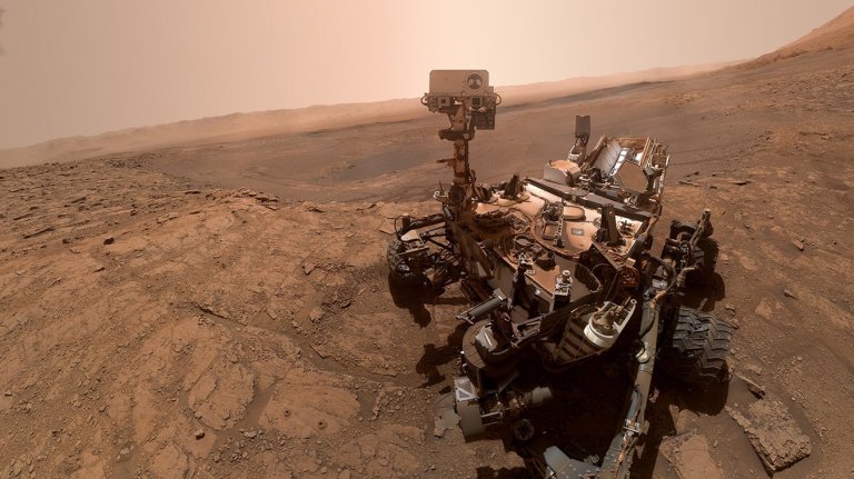 A wide-angle photograph shows a robotic rover positioned on the right side of a dusty, rocky reddish-orange landscape, angled slightly toward the viewer. The detailed vehicle, covered in intricate machinery, thick tires, and a tall camera mast, sits on uneven, cracked ground. In the background, a vast, pale orange sky fades over distant, hazy hills stretching toward the horizon under bright, diffuse sunlight.