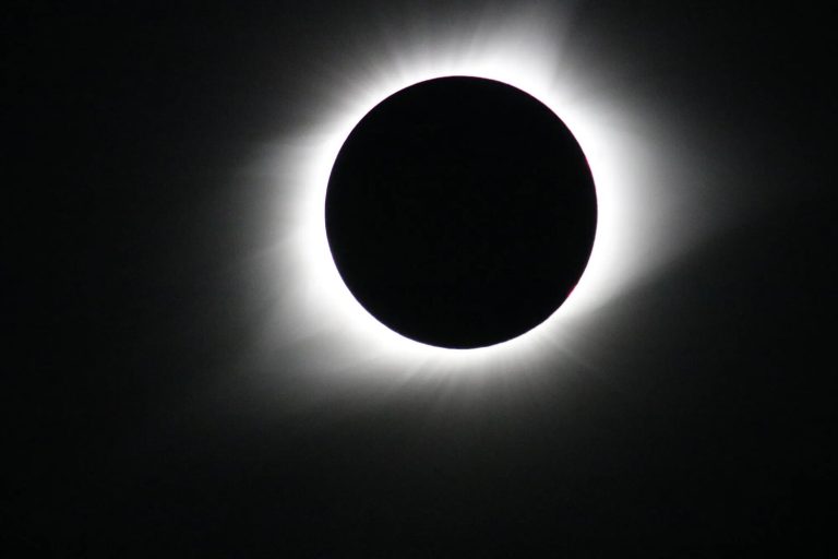 A stark, high-contrast photograph captures a solid black circle perfectly centered against a jet-black background. A bright, glowing ring of white light radiates outward from behind the dark circle, creating a soft, hazy halo with faint streaks extending into the surrounding darkness.