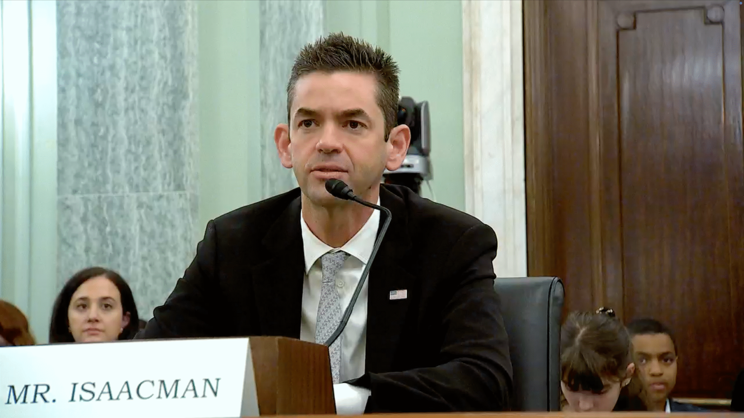 A man in a suit and tie, identified by a nameplate reading "Mr. Isaacman," speaks into a microphone during a formal hearing or panel. Several people sit behind him.