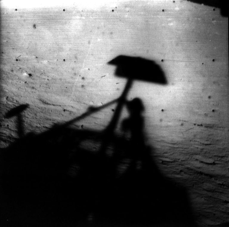 A high-contrast, black-and-white image shows the dark, sharp shadow of a mechanical structure cast against a pale, textured ground surface. The shadow features a prominent, angled central mast topped with a flat, rectangular shape resembling a small awning or panel. The background is a stark, light gray expanse marked with a grid pattern of small, faint dark dots.