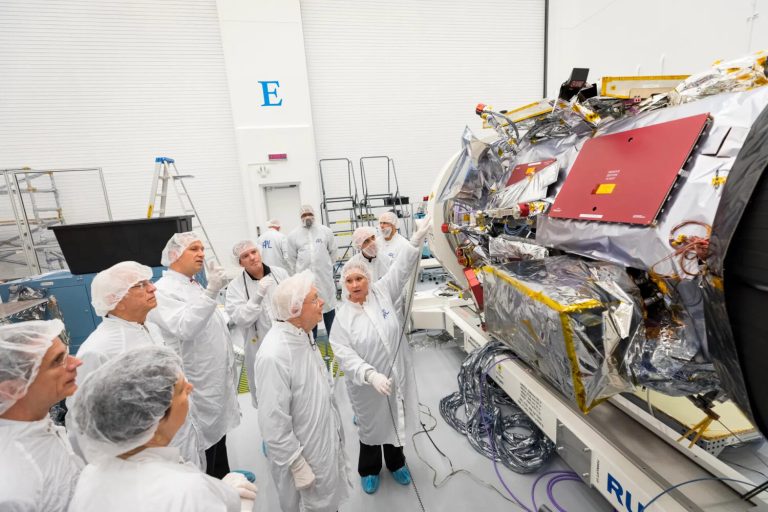 A group of people wearing white protective laboratory suits and hairnets stand in a brightly lit, high-ceilinged room. A woman in the center of the group points toward a large, complex piece of industrial machinery wrapped in shiny silver, gold, and red paneling. The spacious background features white walls, a large blue letter "E," and various ladders and equipment scattered across the gray floor.