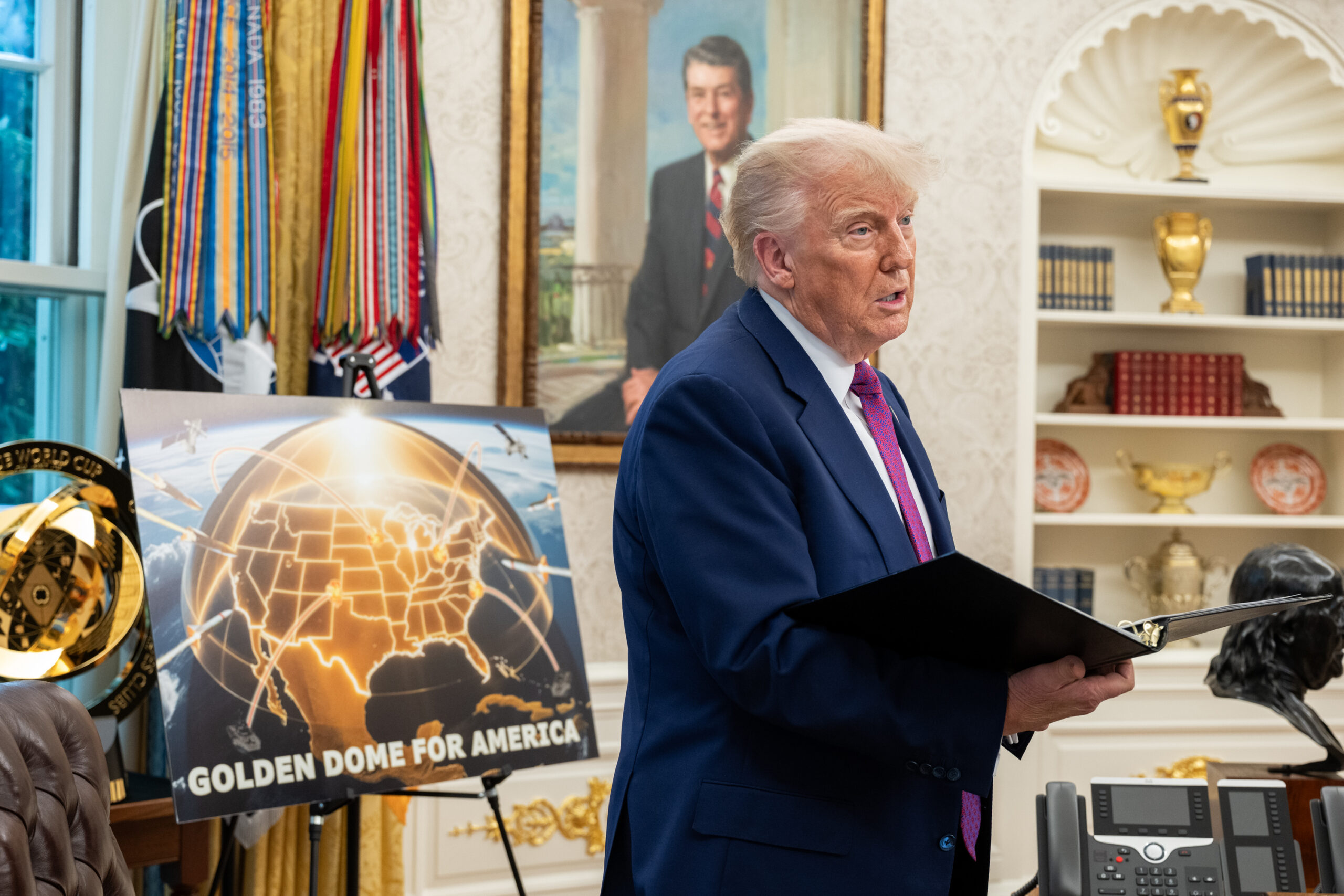 President Donald Trump stands in the Oval Office holding a black folder, speaking near a display board showing a glowing shield-like graphic labeled "GOLDEN DOME FOR AMERICA" over a map of the United States. Behind him are military decorations, a portrait of Ronald Reagan, and shelves with books and golden decorative items.