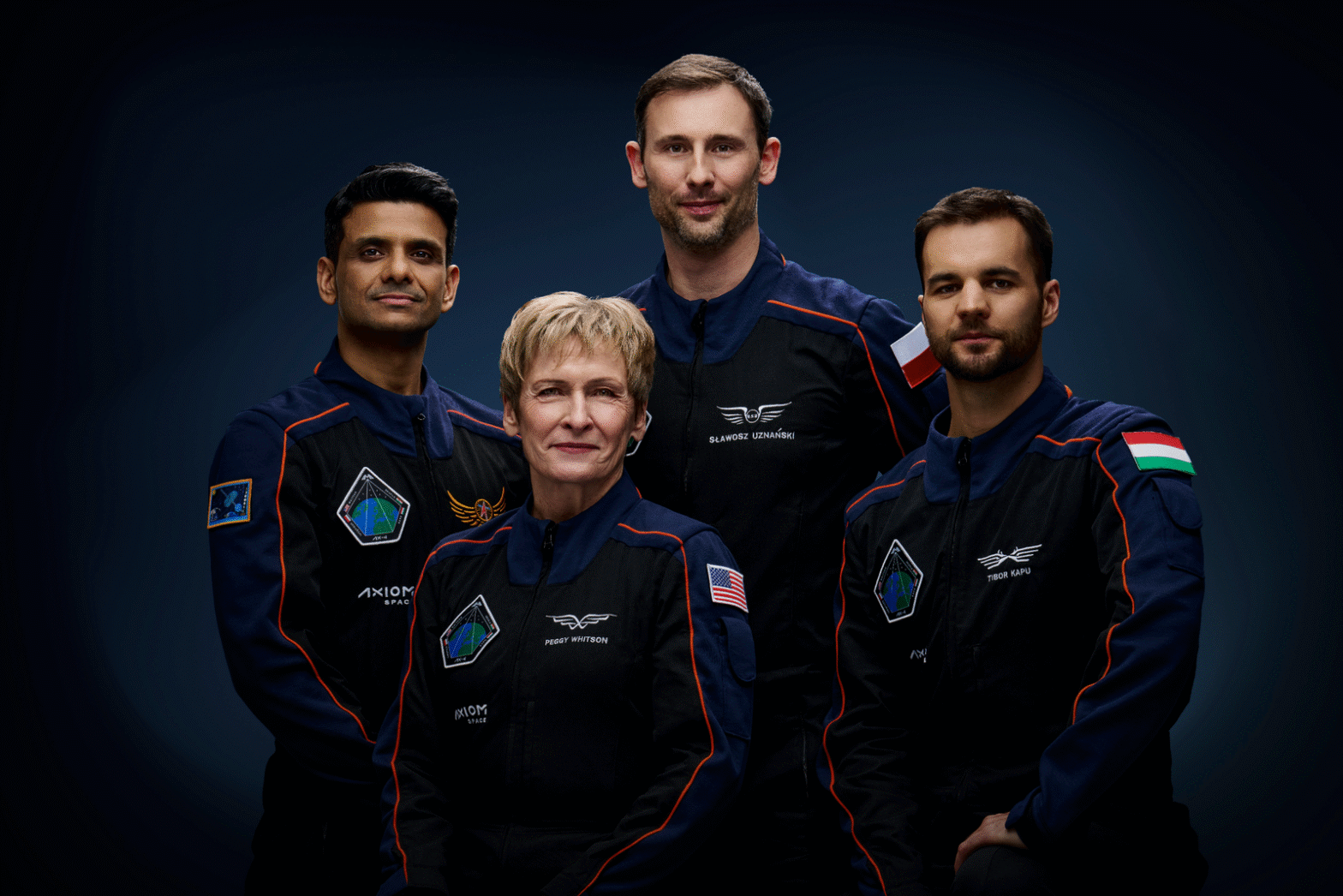 The Axiom Mission 4 (Ax-4) crew poses in their official flight suits against a dark blue background. From left to right: an astronaut from India, Shubhanshu Shukla; NASA astronaut and mission commander Peggy Whitson (front center); European Space Agency astronaut from Poland, Sławosz Uznański (back center); and Hungarian astronaut Tibor Kapu (right). Each crew member wears a navy and black suit with mission patches, national flags, and name tags.