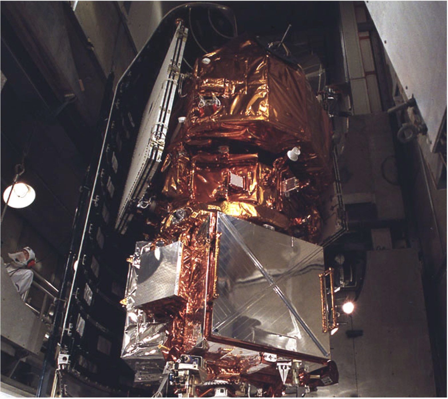 A large, complex satellite stands vertically inside a dimly lit industrial facility, mounted atop a white, cone-shaped rocket component. The upper sections of the satellite are wrapped in crinkled gold and silver metallic foil, accented by flat, dark paneling along one side. In the foreground, a person wearing a white protective suit and mask stands on the right, providing a sense of scale to the brightly illuminated machinery.