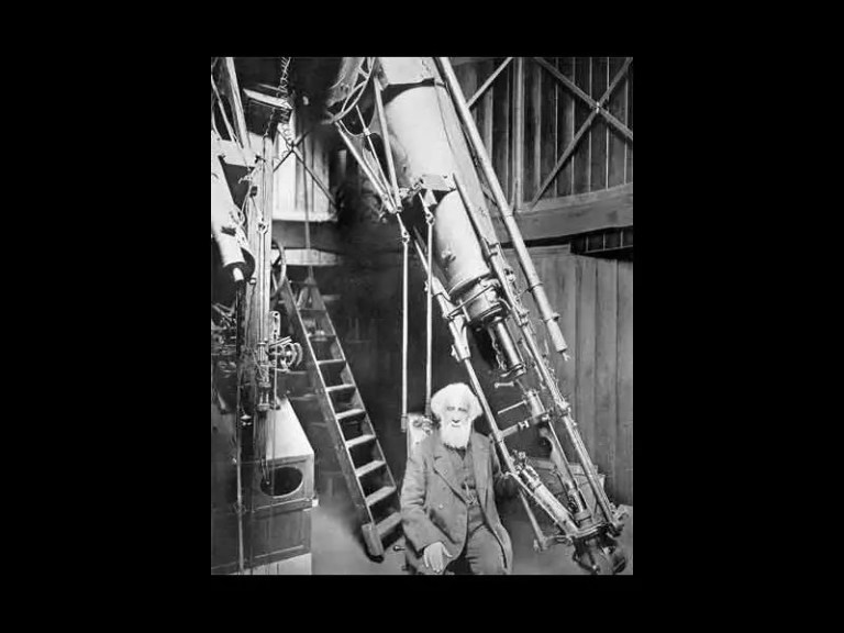 A black-and-white photograph features an elderly man with a long white beard and white hair seated inside a wooden room with high ceilings. He sits directly beneath a massive, metallic telescope tube that slants diagonally down from the upper left toward the lower right of the frame. In the background, a wooden ladder stands to the left of the man against a wall of vertical wooden planks, all under a bright, high-contrast light that illuminates the center of the scene.