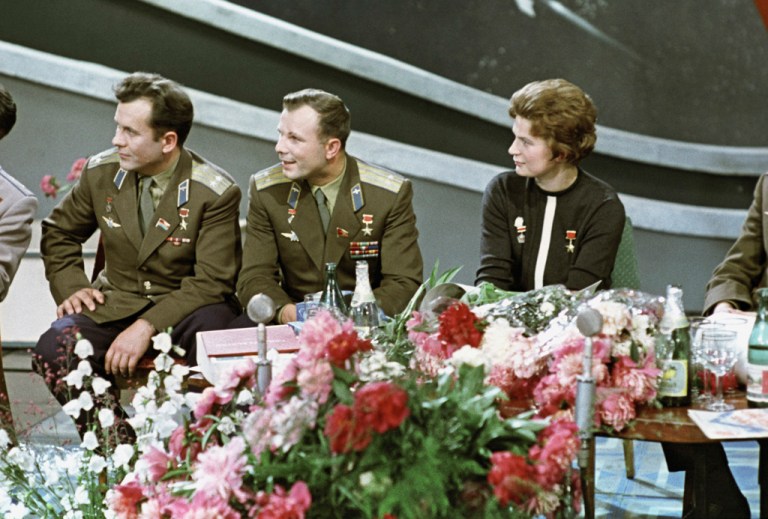 A color photograph shows three people—two men on the left in olive-green military dress uniforms with gold shoulder boards and medals, and a woman on the right in a dark sweater with a white stripe—seated behind a long table covered in large, colorful bouquets of pink and red flowers. The group is captured from the waist up, looking off to the left against a muted gray background with soft, curved horizontal stripes. The scene is brightly lit. All three people sport military medals on their clothing.