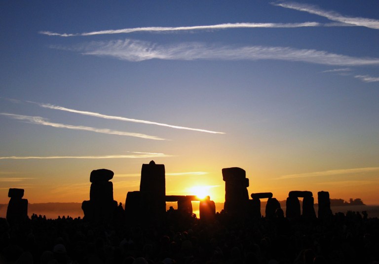 A vibrant sunrise colors the sky with bold streaks of bright pink, purple, and orange behind a silhouette of large stone pillars arranged in a circle. The dark, grass-covered ground in the foreground contrasts sharply with the colorful sky. A crowd of many people stands gathered amongst and around the towering stones, facing toward the bright glow on the horizon.
