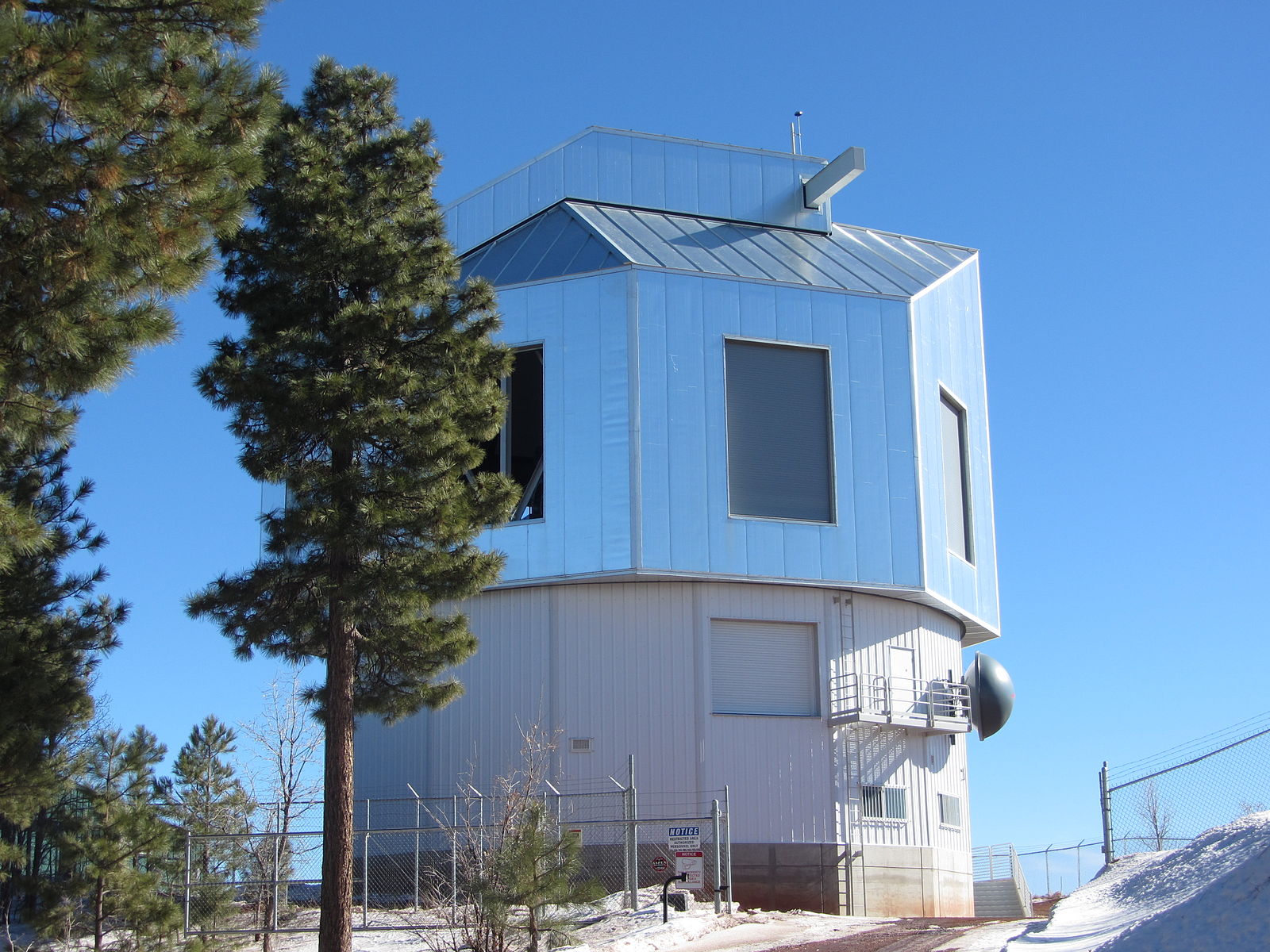 The silver, multi-sided dome of the Discovery Channel Telescope stands under a clear blue sky, surrounded by tall pine trees and patches of snow, with a security fence enclosing the observatory.
