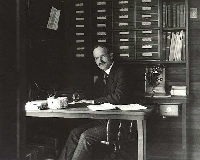 A black-and-white photograph shows a man with a mustache and glasses sitting at a large wooden desk, looking directly at the camera. He wears a dark suit, tie, and striped trousers, with his desk covered in papers and an inkwell. Behind him, a tall wooden cabinet with numerous small drawers and a shelf of books fills the background under high-contrast lighting that highlights his face and the desk surface.