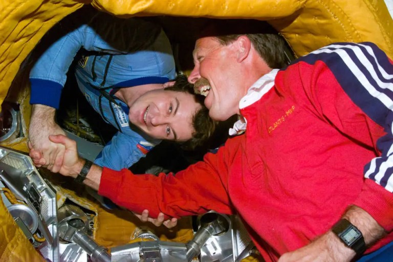 A close-up color photograph captures two smiling men shaking hands inside a cramped, rounded hatchway. The man on the left leans forward in a bright blue jumpsuit, while the man on the right, sporting a mustache, is clad in a vibrant red long-sleeved shirt with white stripes on the shoulder. The entire scene is brightly lit, revealing textured, yellow insulated padding lining the circular tunnel around them.