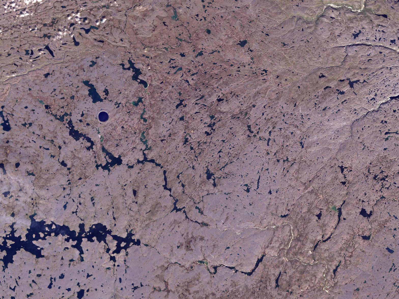 An aerial color photograph shows a vast, textured landscape of brownish-purple terrain crisscrossed by numerous small, dark blue lakes and winding rivers. In the upper-left section of the frame, a distinct, perfectly circular deep blue lake stands out from the surrounding irregular waterways. The entire scene is evenly lit, emphasizing the intricate, patterned network of water pockets scattered across the dry land.
