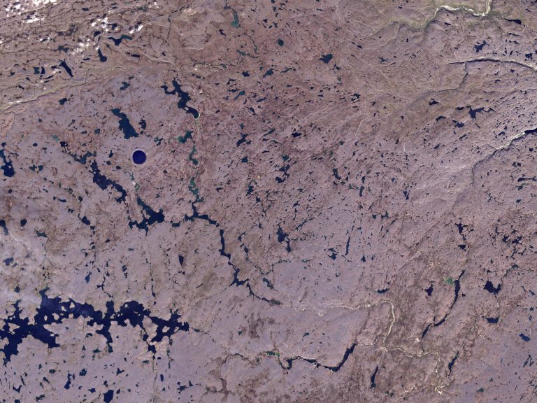 An aerial color photograph shows a vast, textured landscape of brownish-purple terrain crisscrossed by numerous small, dark blue lakes and winding rivers. In the upper-left section of the frame, a distinct, perfectly circular deep blue lake stands out from the surrounding irregular waterways. The entire scene is evenly lit, emphasizing the intricate, patterned network of water pockets scattered across the dry land.