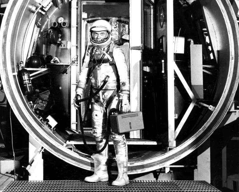 A black-and-white photograph shows a person in a gleaming, metallic space suit and helmet standing inside a large, circular opening of a technical structure. The individual stands centrally, holding a small rectangular case in one hand and a hose in the other, positioned on a metal grate floor. The scene is brightly lit with sharp contrast, highlighting the reflective surfaces of the suit against a complex background of machinery, frames, and deep shadows.