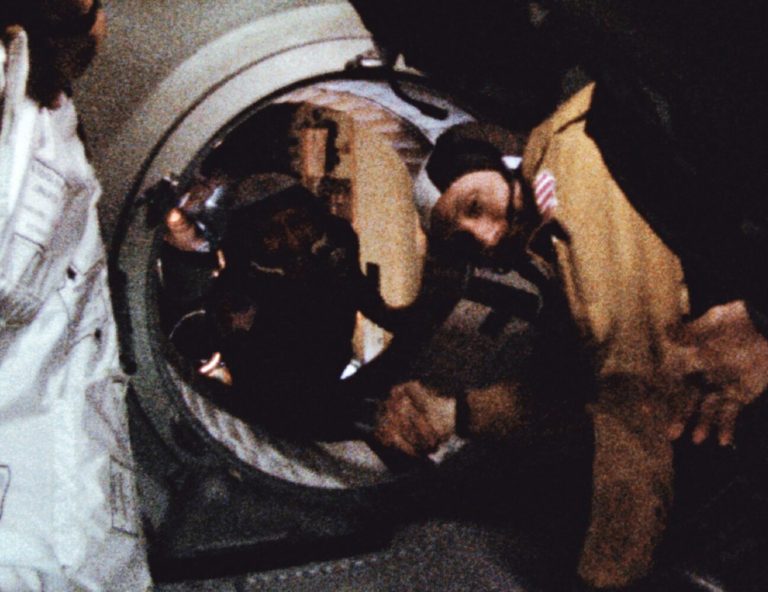 Two people are shaking hands through a circular hatch that connects two dark, enclosed spaces. The person on the right wears a tan jacket, while the other is partially visible inside the opening. The scene features high contrast, with deep shadows surrounding the brightly lit hands and faces.