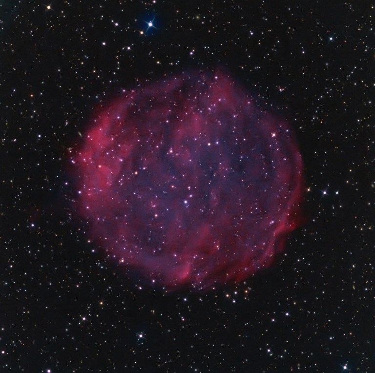 A spherical, faintly glowing pink nebula known as Purgathofer-Weinberger 1 floats against a dark star-filled background. The nebula’s diffuse, circular shape appears semi-transparent, with a mottled texture of filamentary gas and scattered stars shining through and around it. The outer edges glow slightly brighter, forming a faint shell-like structure. Countless distant stars twinkle across the image.