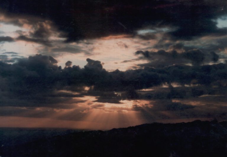 A wide landscape color photograph captures a dramatic sky filled with thick, dark gray clouds layered across the frame. Sunbeams break through gaps in the clouds, casting fans of dim orange and white light downward toward a silhouetted, dark horizon line. The scene is heavy with shadow, creating a deep contrast between the dark cloud formations and the glowing pockets of twilight sky.