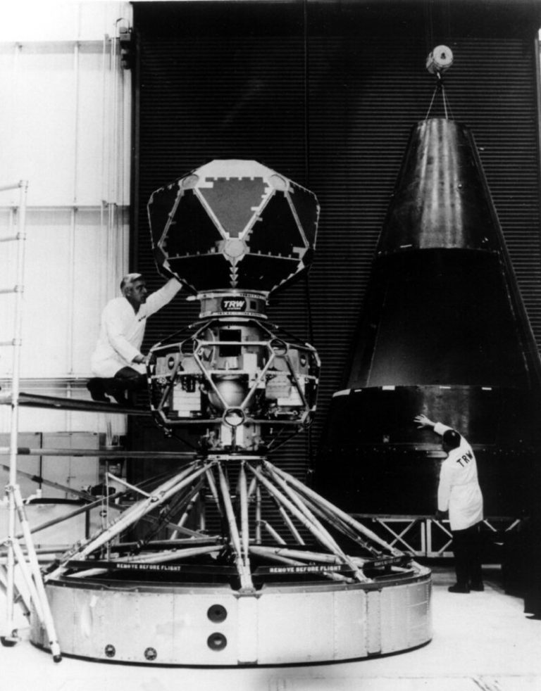 A black-and-white photograph shows a large, complex piece of machinery in an industrial setting alongside a tall, cone-shaped capsule suspended from above. Two men in white lab coats work on the equipment: One sits on a scaffolding platform to the left adjusting a multi-sided geometric sphere atop the machinery, while the other stands on the floor to the right touching the dark cone. The scene is evenly lit with clear contrast, set against a background of large, dark corrugated bay doors and workshop scaffolding.