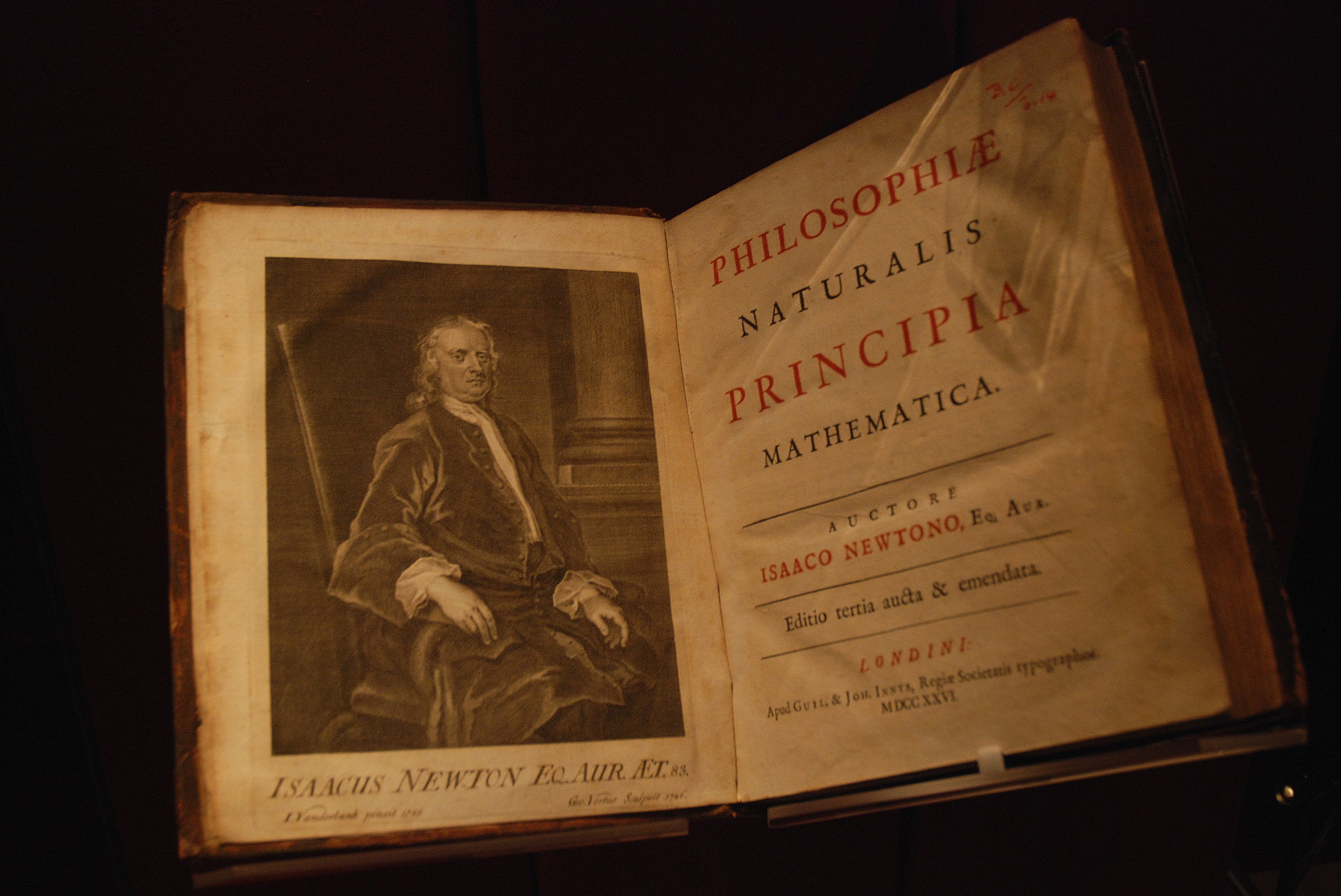 An open, antique book fills the frame against a dark background. The left page features a black-and-white illustrated portrait of a seated man in 18th-century attire, while the right page shows a title layout with large red and black Latin text on aged, off-white paper. Dim, warm lighting illuminates the book from above, creating a strong contrast with the deep shadows around it.