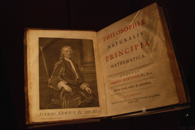 An open, antique book fills the frame against a dark background. The left page features a black-and-white illustrated portrait of a seated man in 18th-century attire, while the right page shows a title layout with large red and black Latin text on aged, off-white paper. Dim, warm lighting illuminates the book from above, creating a strong contrast with the deep shadows around it.