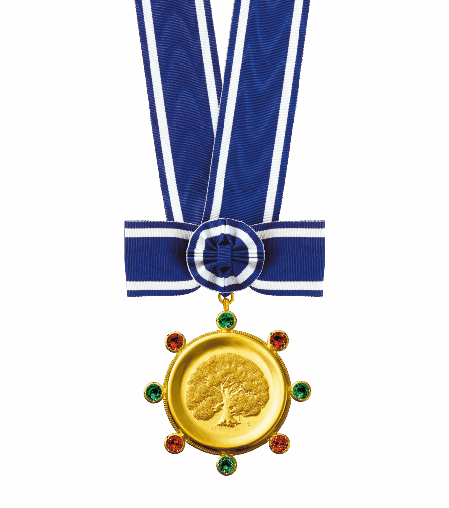 A color photograph shows a gold medal hanging from a blue-and-white striped ribbon, centered against a solid white background. The circular, textured gold medal features an engraved tree in the middle and is adorned with alternating red and green gems around its outer edge. The ribbon above it is arranged into a neat bow with a circular pleated rosette at the center.