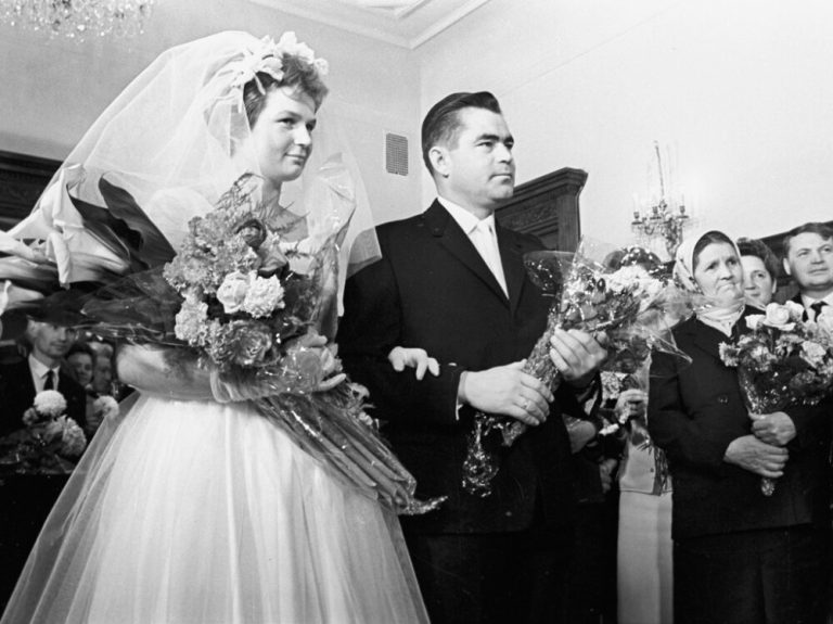 A black-and-white photograph shows a bride and groom standing together in the center of the frame, both holding large bouquets of flowers wrapped in clear cellophane. The bride wears a white gown with a full tulle skirt, a matching veil with a floral crown, and white gloves, while the groom wears a dark, tailored suit with a white shirt and tie. They stand arm-in-arm, looking off-camera to the right, under bright indoor lighting with several onlookers visible in the background.