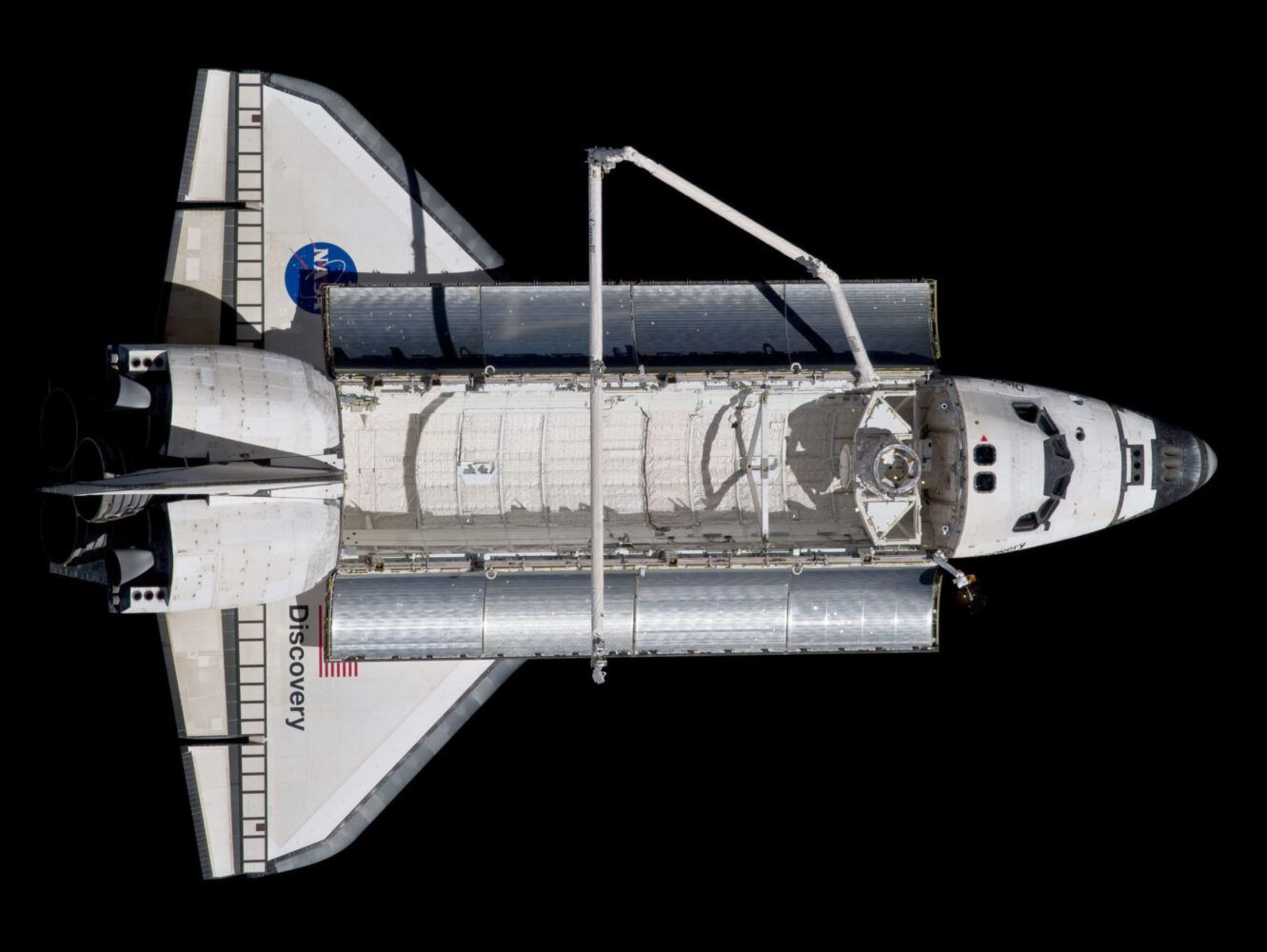 An overhead view shows a white and grey space shuttle floating horizontally in the center of the frame against a solid black background. Its large, open cargo bay doors reveal a textured, cylindrical white module inside, with a long, thin robotic arm extending over the bay. Bright, direct sunlight from the upper right casts deep shadows across the shuttle’s body, highlighting the details of its wings, cockpit windows, and rear engines.