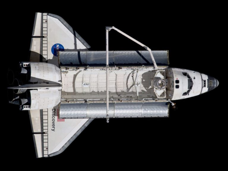 An overhead view shows a white and grey space shuttle floating horizontally in the center of the frame against a solid black background. Its large, open cargo bay doors reveal a textured, cylindrical white module inside, with a long, thin robotic arm extending over the bay. Bright, direct sunlight from the upper right casts deep shadows across the shuttle’s body, highlighting the details of its wings, cockpit windows, and rear engines.