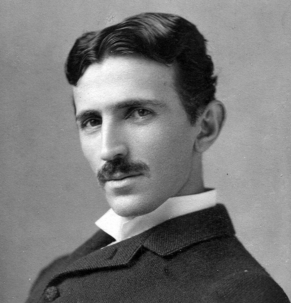 A black-and-white studio portrait shows a man with dark, neatly parted hair and a thick mustache looking directly forward. He is dressed in a dark, textured jacket with a high white collar visible at his neck. The lighting is soft and even across his face, set against a plain, light grey background.