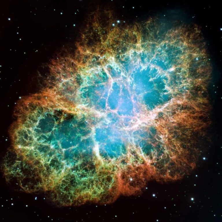 A large, oval-shaped cloud of colorful gas expands outward across the frame against a black background dotted with tiny stars. The center of the cloud glows with a bright, hazy blue-white light, which transitions into an intricate web of yellow, green, and fiery red filaments weaving along the outer edges. There is sharp contrast between the vibrant, glowing gases of the central structure and the dark void of space surrounding it.