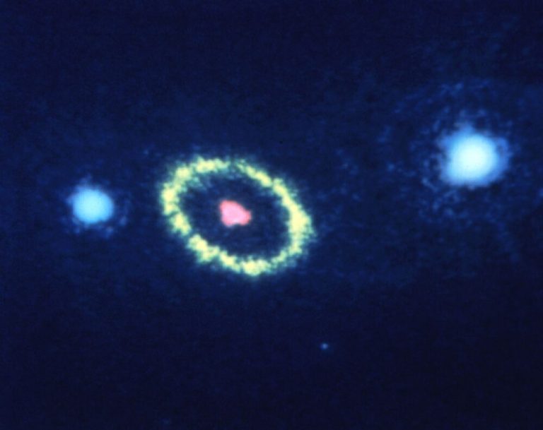 A grainy astronomical photograph shows three bright glowing shapes arranged in a slight horizontal arc across a dark blue background. At the center, a fuzzy, pale yellow oval ring encircles a small, bright red dot. Flanking the ringed shape on either side are two bright, soft-edged blue-white spheres, with high contrast separating the luminous central objects from the surrounding dark expanse.