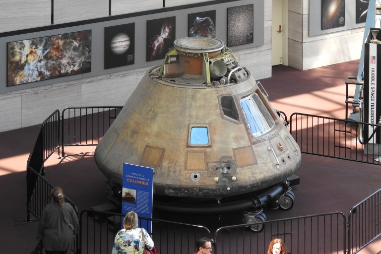 A high-angle photograph shows a cone-shaped space capsule resting on a wheeled platform in the center of a museum hall, surrounded by black metal barricades. The capsule's metallic, tarnished exterior displays shades of gold, brown, and grey with a bright blue reflection visible in one of its small square windows. Bright sunlight streams across the carpeted floor, while visitors view the display from the foreground and framed space photographs hang on the grey stone wall in the background.
