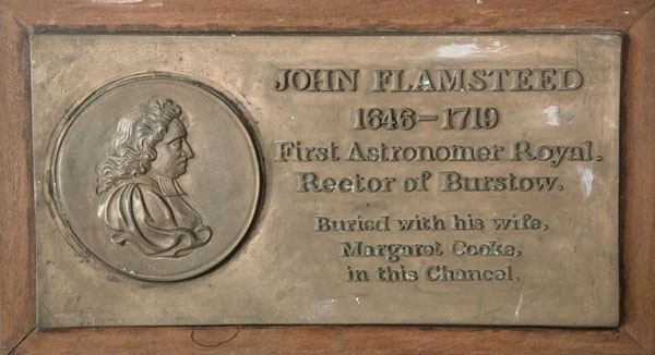 A rectangular bronze wall plaque with a aged metallic patina is set inside a dark wooden frame. On the left side, a circular relief medallion features a profile bust of a man in historical attire, while raised lettering on the right displays the name "JOHN FLAMSTEED," dates, titles, and a memorial inscription. Even lighting across the metallic surface highlights the raised details against the smooth, tarnished brass background.