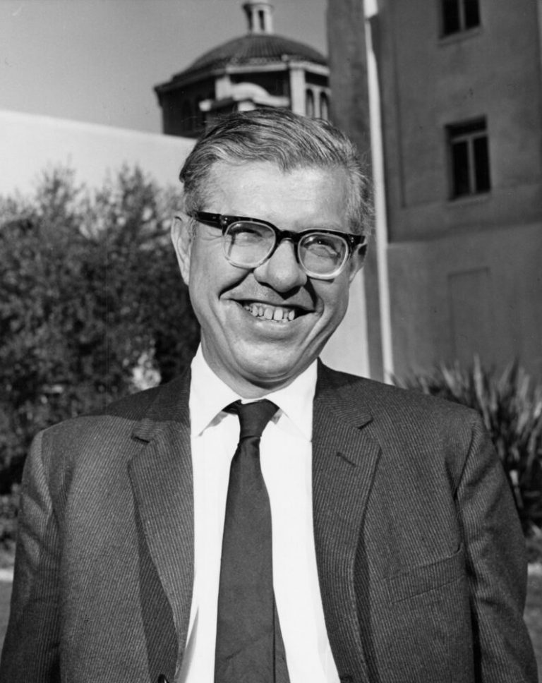 A black-and-white portrait shows a man wearing thick-rimmed glasses, a dark suit jacket, and a tie, smiling broadly in the center of the frame. Direct daylight illuminates his face and the front of his white shirt, creating clear contrast against his dark jacket. In the blurred background, trees and a multi-story building featuring a domed tower stand under a bright sky.