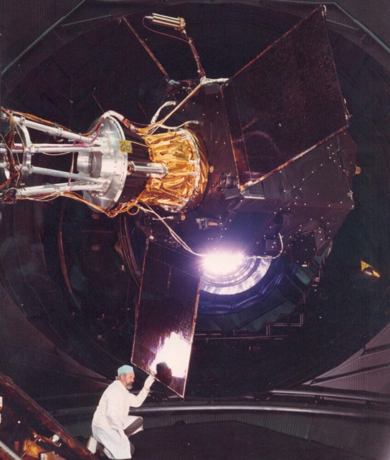 A complex, octagonal satellite wrapped in black panels and gold foil dominates the upper portion of the frame inside a large, dark circular testing chamber. Near the bottom left, a person wearing a white lab coat and blue surgical cap kneels while touching one of the satellite’s rectangular panels. A intense white light shines from the center of the structure, creating sharp highlights and high contrast against the dark background.