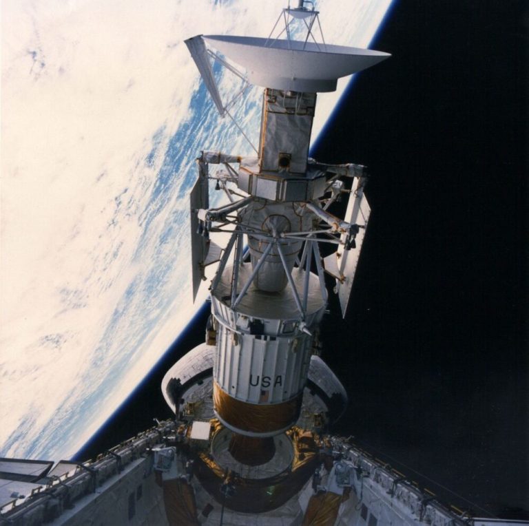 A tall, white spacecraft topped with a large dish antenna extends vertically through the center of the frame above an open cargo bay. The background is sharply divided, with the bright, curved blue-and-white surface of Earth filling the upper left and the deep black of space on the right. Direct sunlight brightly illuminates the vehicle's metallic gold foil and white structural panels, creating high contrast against the dark void.
