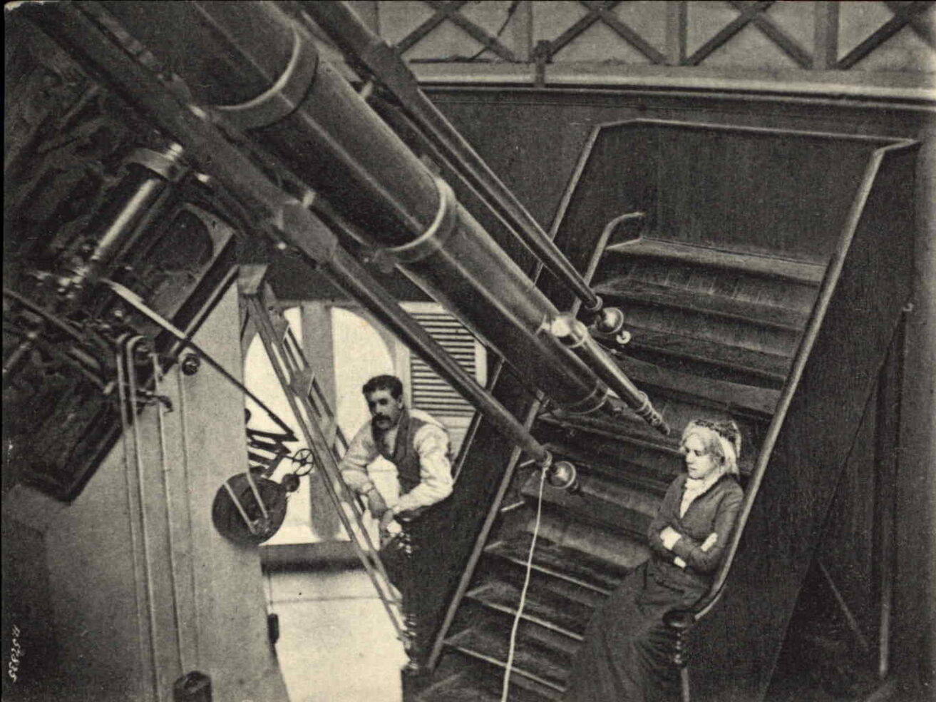 A vintage sepia-toned photograph shows a large, long telescope extending diagonally across the left side of the frame. On the right, a woman sits on a set of wooden stairs near the eyepiece end of the telescope, while a man with a mustache leans against a ladder in the background. The indoor space is softly lit, with muted contrast highlighting the wooden structure, metal equipment, and dark clothing of the two figures.