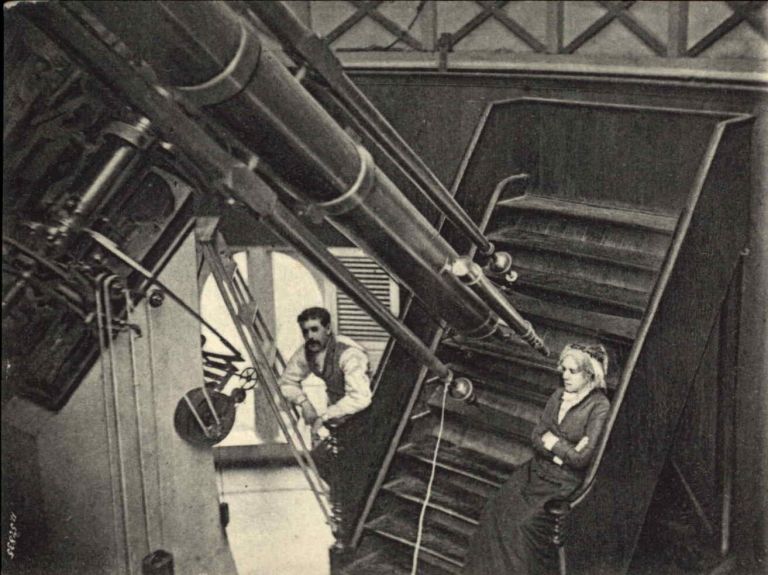 A vintage sepia-toned photograph shows a large, long telescope extending diagonally across the left side of the frame. On the right, a woman sits on a set of wooden stairs near the eyepiece end of the telescope, while a man with a mustache leans against a ladder in the background. The indoor space is softly lit, with muted contrast highlighting the wooden structure, metal equipment, and dark clothing of the two figures.