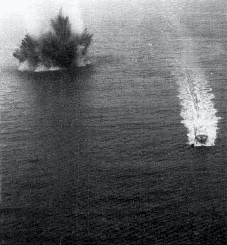 A high-angle, black-and-white photograph shows a large plume of water bursting upward from an explosion in the upper left section of the open sea. On the right side, a small boat moves away from the splash, leaving a long, bright white wake across the dark, rippling water. The image features strong contrast between the dark grey ocean surface and the pale, frothy water from both the blast and the boat's trail.