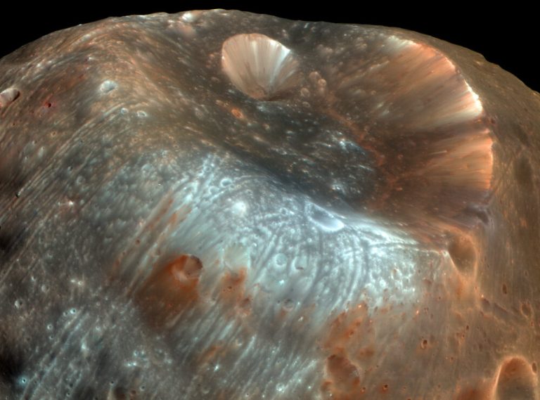 A close-up photograph shows the dusty, cratered surface of a moon filling most of the frame against a black space background. A bowl-shaped crater dominates the upper right, featuring steep walls lined with reddish-brown and pale tan streaks. A bright, pale blue-grey glow highlights the textured, grooved terrain surrounding smaller craters across the center and lower-left regions.