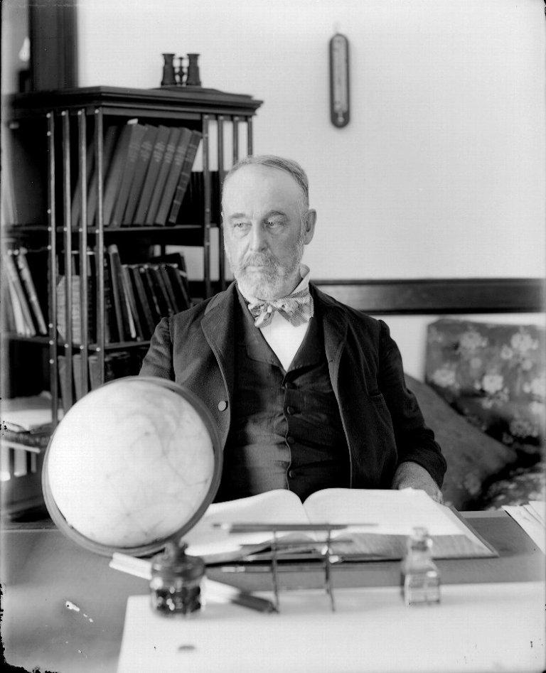 A black-and-white portrait shows a bearded man sitting at a dark wooden desk in the center of the frame, facing slightly to the left. On his desk in the foreground are an open book, a small globe, inkwells, and writing utensils. Behind him, a bookcase filled with books stands on the left, while a plain wall with a small thermometer and a floral-patterned sofa complete the background.