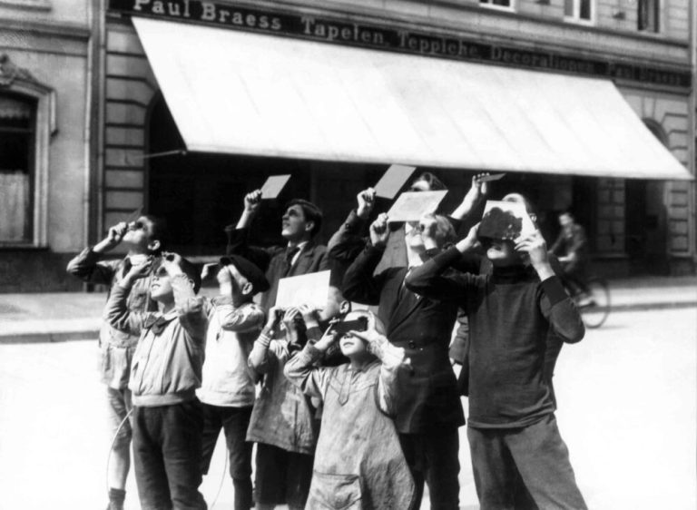 A vintage black-and-white photograph captures a crowd of adults and children standing gathered outdoors, looking up toward the sky. Most individuals hold small rectangular cards, dark glass plates, or paper up to their eyes to shield their vision. Bright daylight illuminates the figures in the foreground, contrasting against the darker storefront with a large white awning and signage in the background.