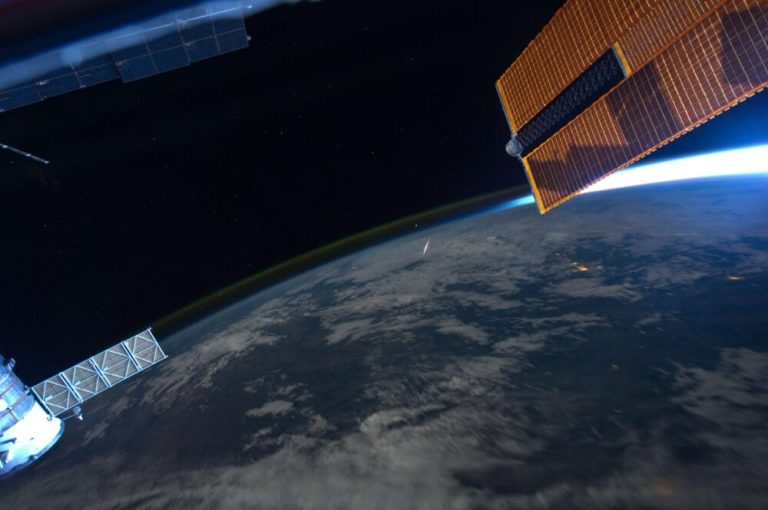 An orbital photograph shows the curved, cloud-covered surface of Earth stretching diagonally across the lower half of the frame under a pitch-black sky. A thin, glowing green band outlines Earth's horizon, while a bright streak of light flashes over the atmosphere near the center. In the upper right and lower left corners, metallic spacecraft structures and solar panel arrays extend into the frame, lit brightly against the dark expanse of space.