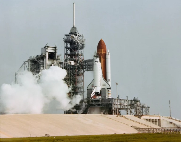 A white space shuttle attached to a large rust-orange fuel tank and white booster rockets stands upright on a launch pad next to a tall, grey metal service tower. A billowing plume of thick white smoke erupts from the base of the tower on the left, contrasting against the pale grey sky background. Daytime lighting brightly illuminates the shuttle and the tan, sloped concrete ramp leading up to the launch platform in the foreground.